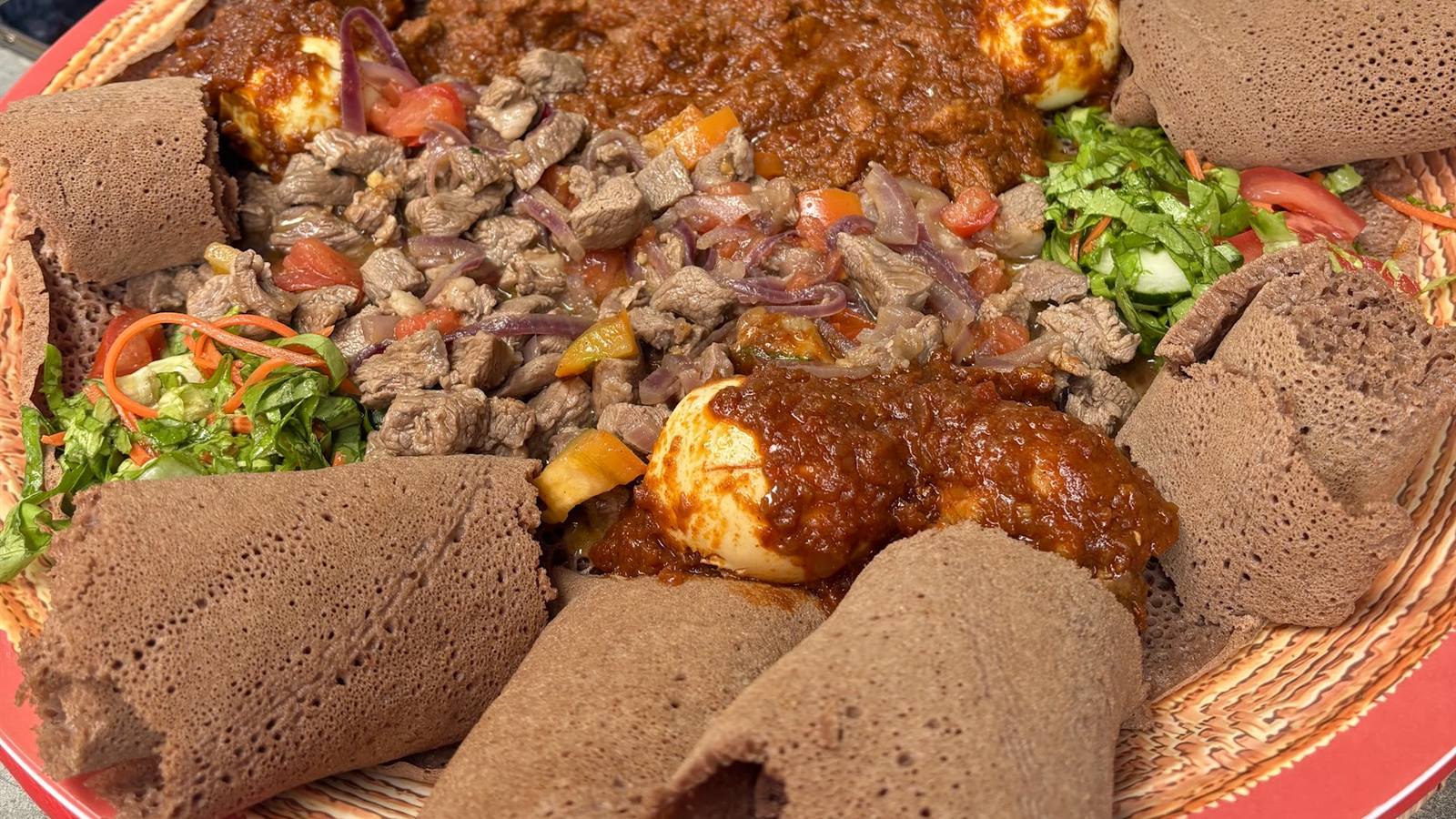 Een grote injera-schotel met stoofpotjes van rundvlees, kip, ei en verse salade, met opgerolde injera langs de rand.
